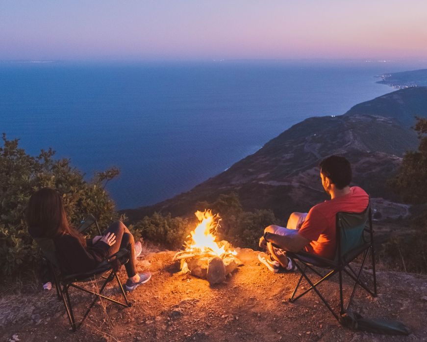 Two people sit around a campfire on a hillside overlooking the ocean at dusk. (Photo: seyfi durmaz, Pexels)