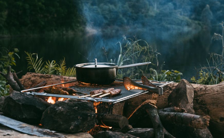 Campfire cooking beside a lake, with a pot and food grilling over an open flame surrounded by rocks.