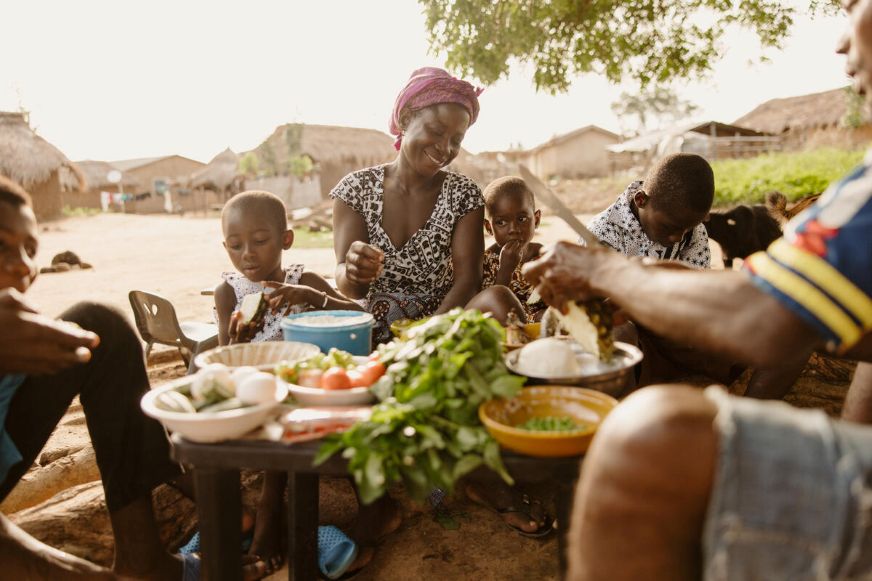 Family members prepare and share a meal outdoors, surrounded by fresh vegetables and ingredients, in a rural community setting. (Ghana, 2025)