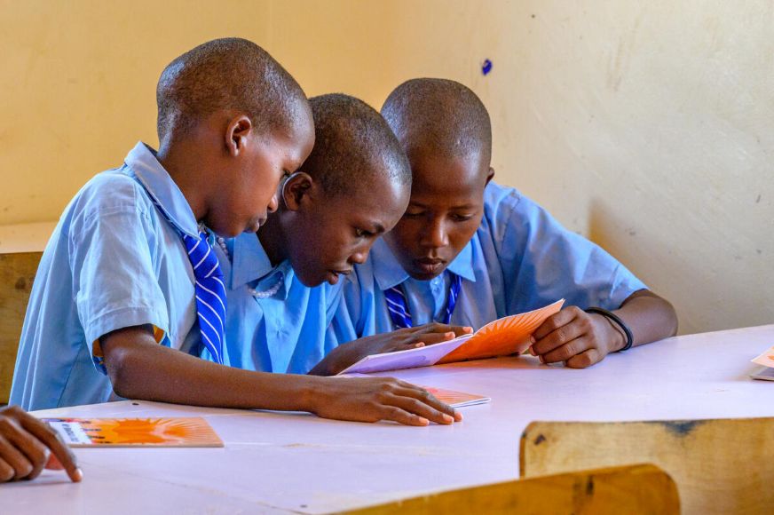Three students in school uniforms sit at a classroom table reading and studying from a book together. (Kenya, 2026)