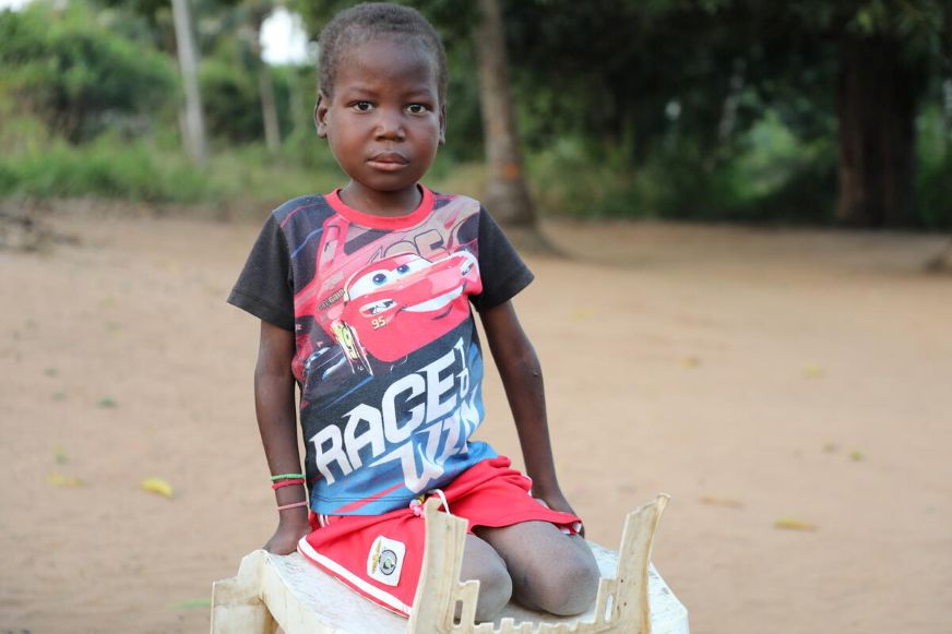 Clementino sits outdoors on a wooden structure wearing a red and black T-shirt and red shorts. (Mozambique, 2024)