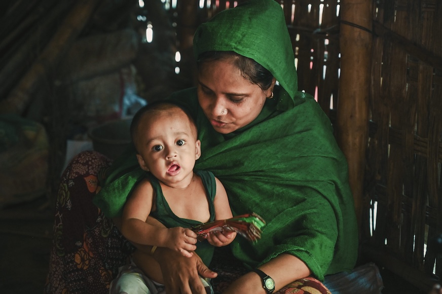 In Bangladesh, a woman in a vibrant green shawl sits on the ground with an infant playing in her lap.