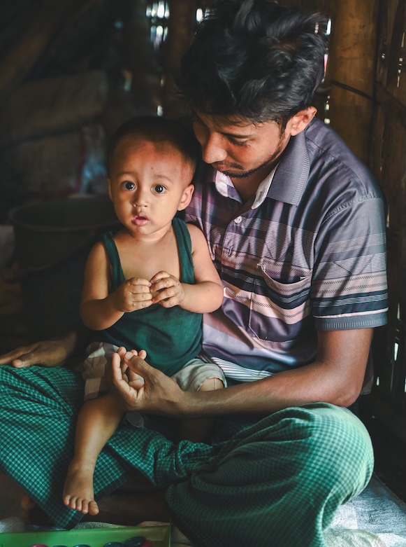 In Bangladesh, a man wearing a purple polo shirt and green pants cradles his infant son, who looks at the camera.