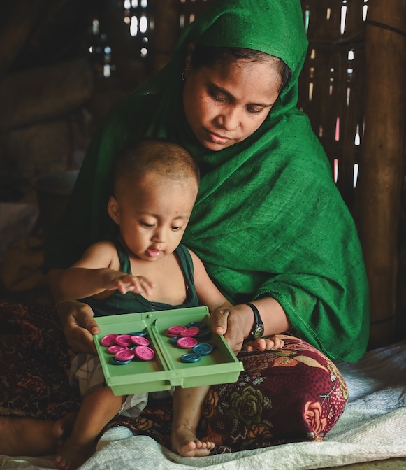 In Bangladesh, a woman in a vibrant green shawl sits on the ground with an infant playing in her lap.