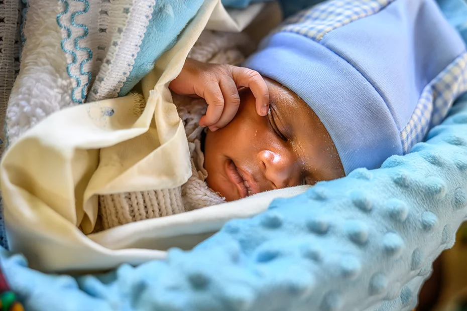 A swaddled newborn baby sleeps with his hand touching his face.