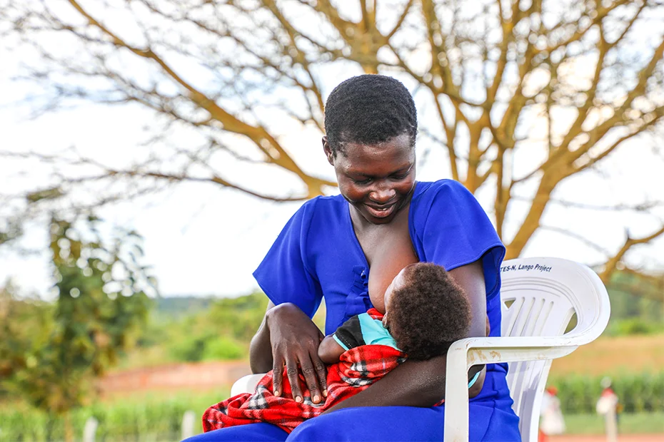A mother sits outdoors in a vibrant blue dress, cradling her baby as she breastfeeds.
