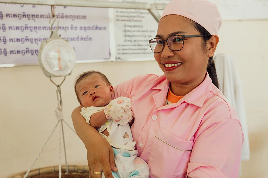 A cheerful midwife holds a newborn baby, preparing to weigh it on a scale.