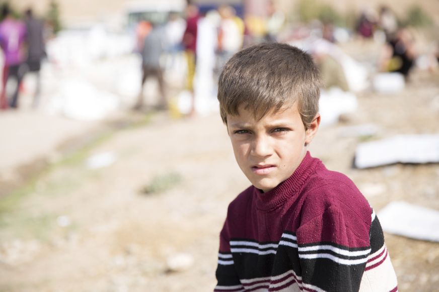 A school-aged boy looks toward camera, in front of a rough, outdoor area.
