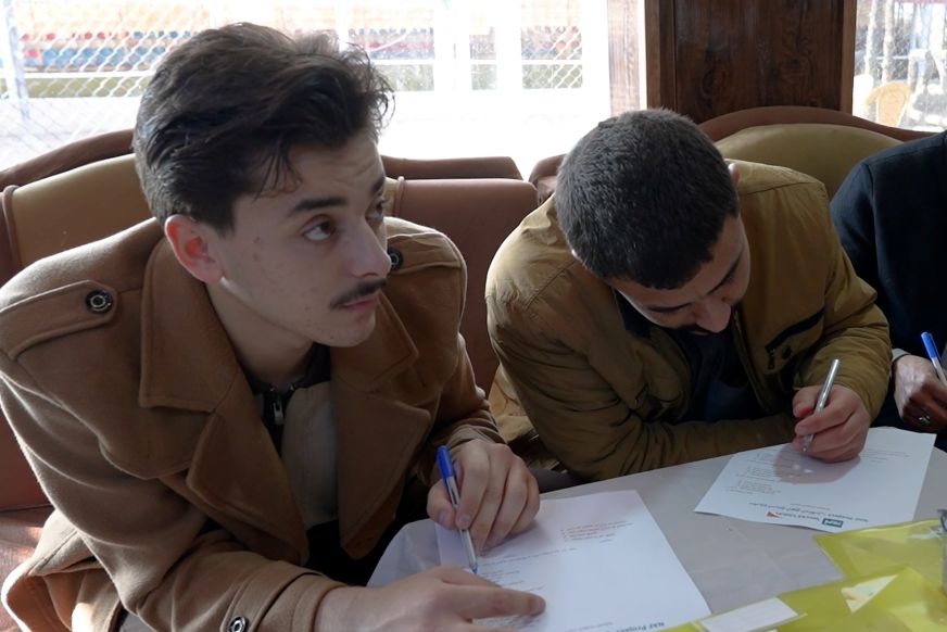 Two young men write on papers on a table, as one looks up and off-camera.