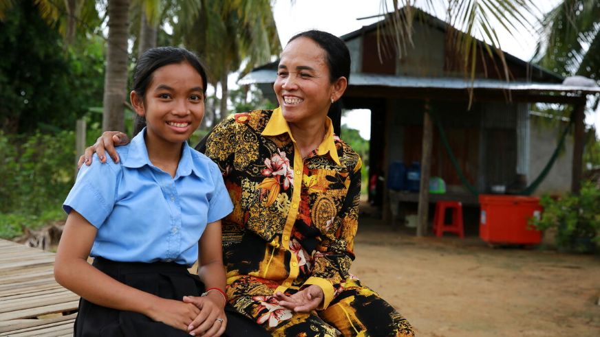 Chandy, in her school uniform, sits smiling with her mom outside their home in Cambodia.