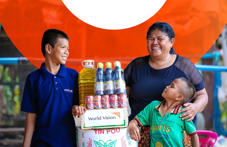 Two boys and a woman standing beside a stack of packaged food items, smiling.