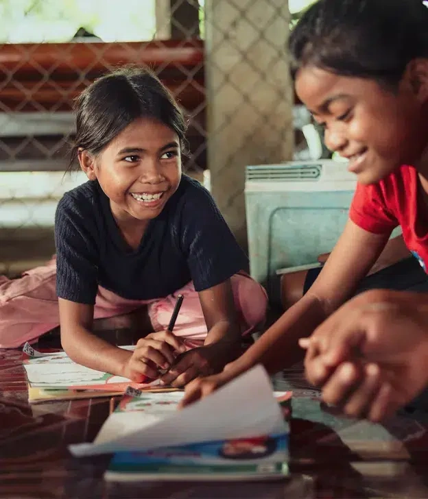 Children sit on the floor together, drawing and writing during a group learning activity indoors.