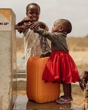 Two small children collect water from a hand pump while an adult sits nearby at a community water point.
