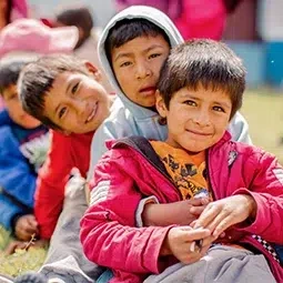 A group of children sit in a line on the grass during an outdoor group activity.