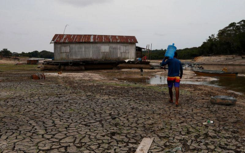 A man walks across dried ground, carrying a water bottle on his shoulder.