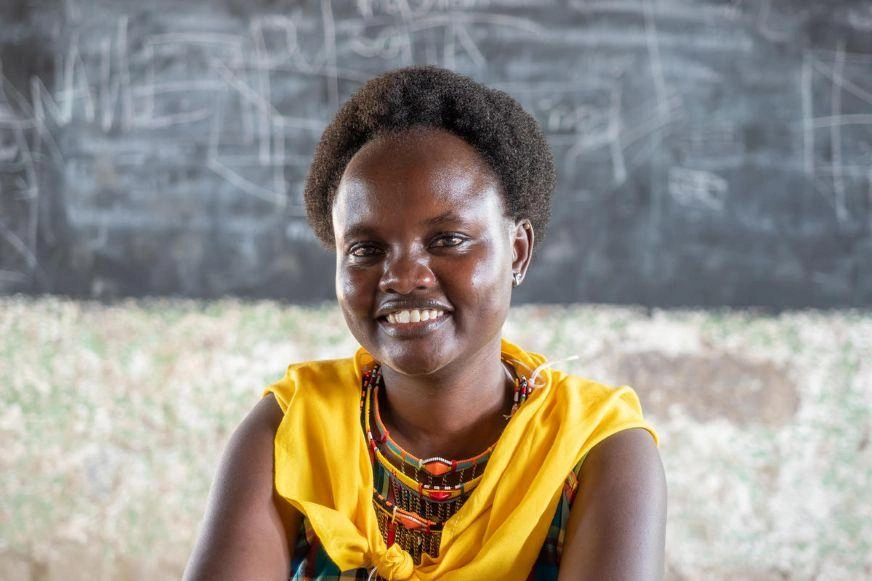 A young woman draped in a yellow scarf sits in front of a chalkboard covered with writing. (Kenya, 2018)