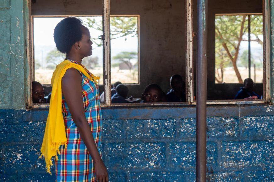 A woman in a yellow shawl and blue patterned dress stands beside a classroom wall, looking through a window at the students inside. (Kenya, 2018)
