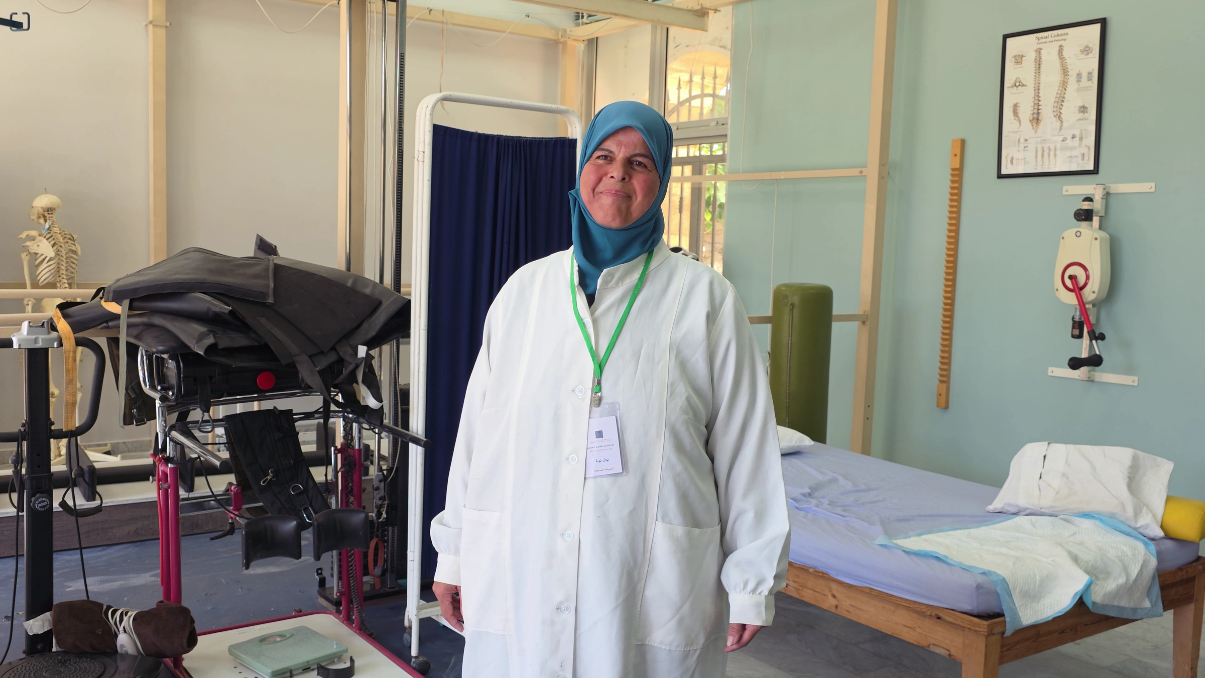 Nawal, a nurse, is wearing a lab coat and lanyard as she stands inside a room at the Derdghaya Socio-Medical Centre