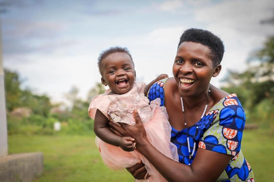 A smiling woman wearing a blue patterned top holds a laughing baby outdoors in a grassy setting. (Uganda, 2024)
