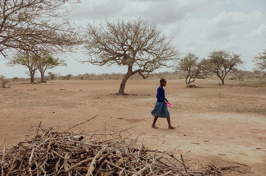 A girl carries an empty dish while walking on a dusty path in an arid landscape.