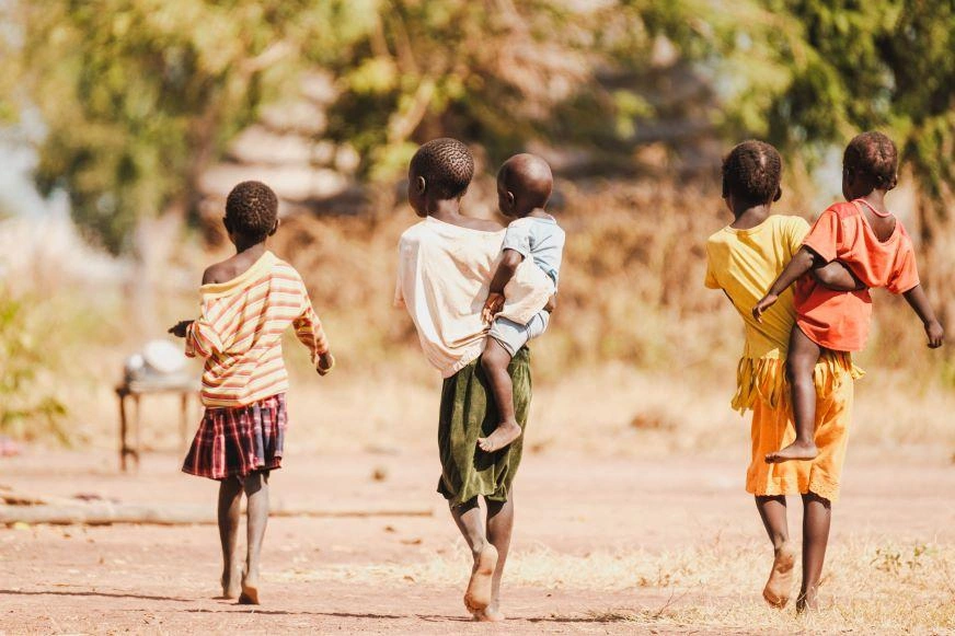 Three children walk on a dirt road in South Sudan. Two have toddlers on their hips.