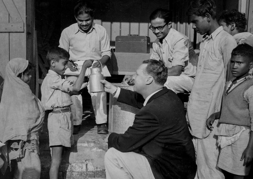 In a black and white image, a boy holds a metal milk container up while a man in a black suit jacket talks to him.