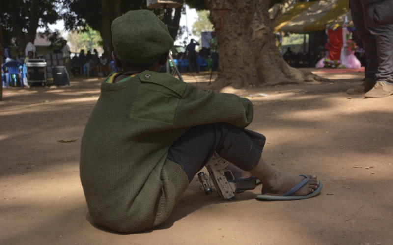 A young boy sits on the ground outside, holding a gun.