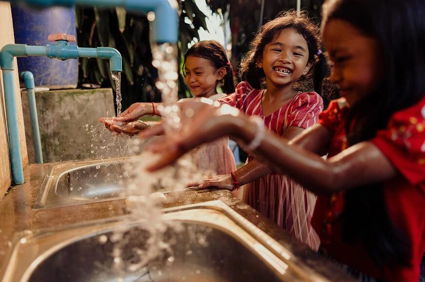 Three young girls laugh and smile while washing their hands under clean, running water at a row of outdoor sinks. The sunlight highlights the splashing water as