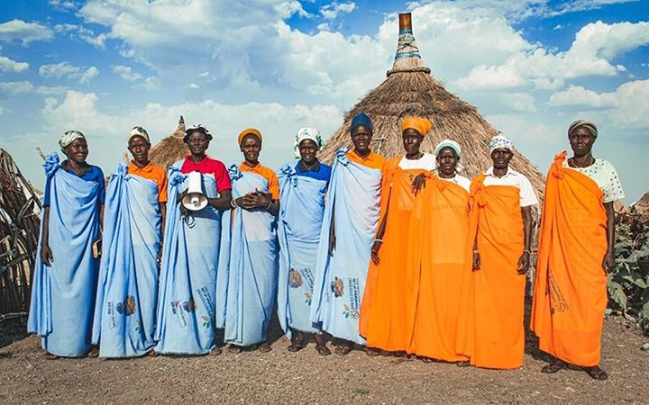 A row of women in brightly coloured robes and headscarves, stand facing the camera. One woman holds a megaphone.