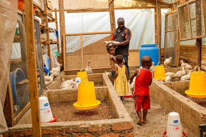 Small children and their father tend chickens inside a light-filled structure.