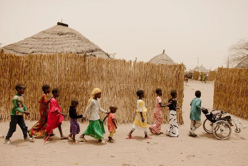 A young boy pushes a child in a wheelchair, while nine children follow in a line behind them.