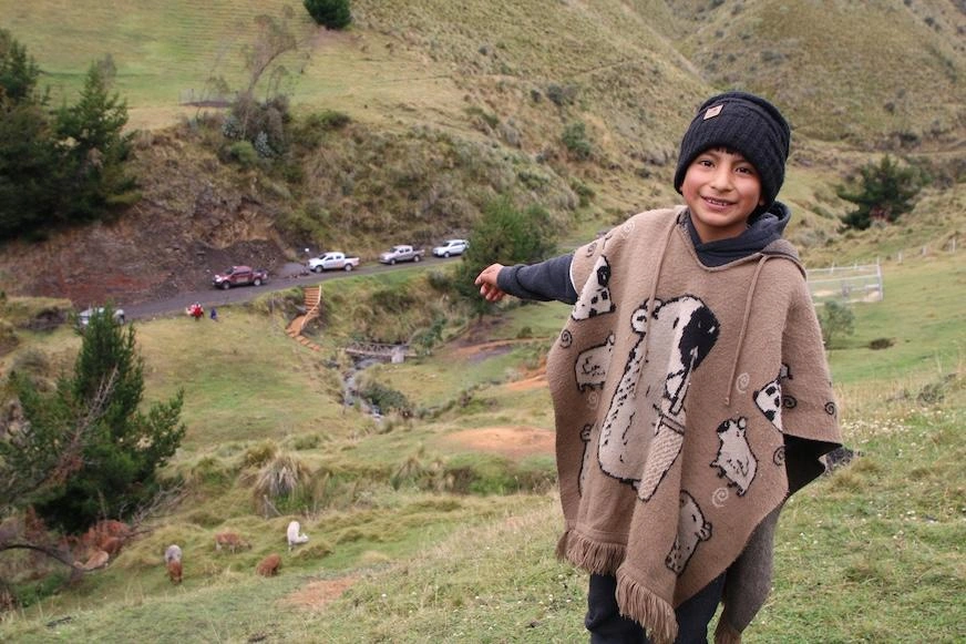 A young boy wearing a winter hat and knit poncho gestures to alpacas on the hill below him.