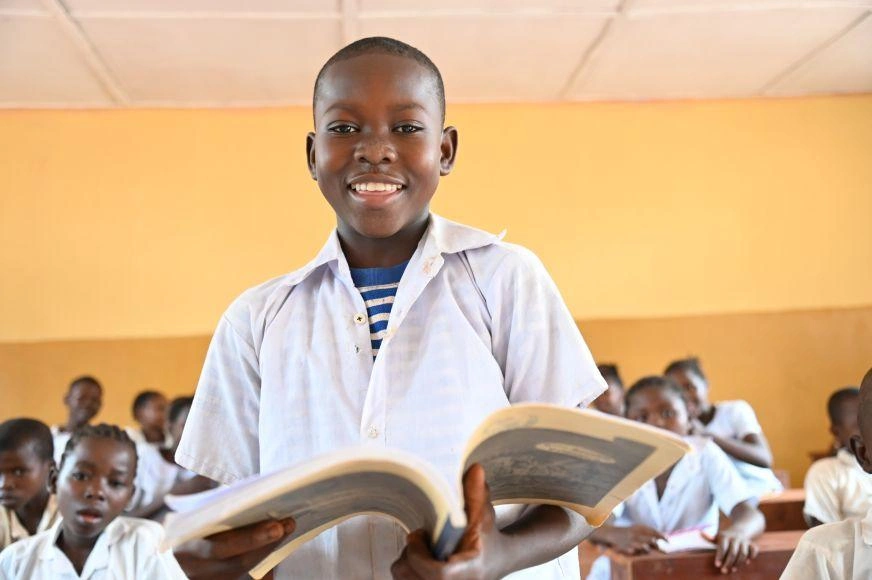 An adolescent boy in the Democratic Republic of Congo stands smiling in a yellow-walled classroom, holding an open book.