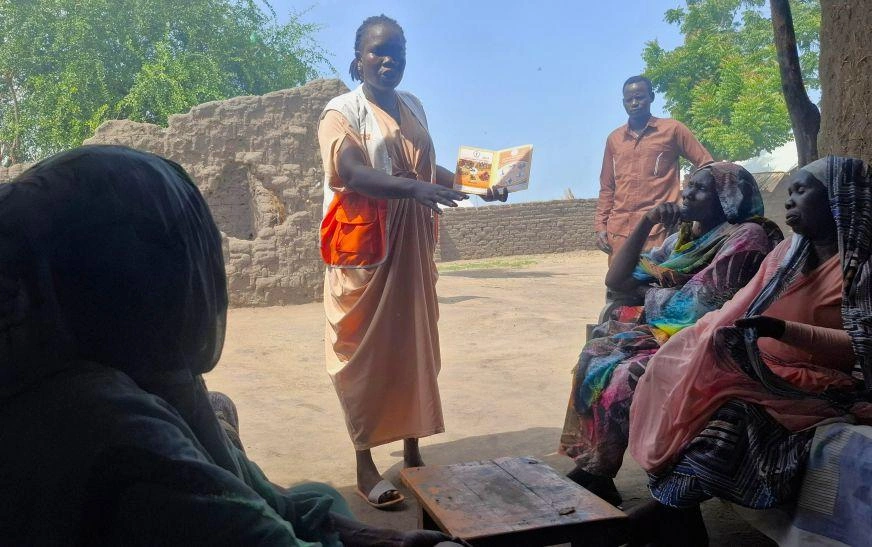 In Joda, South Sudan, a woman wearing an orange World Vision vest stands in front of a group of people presenting hygiene education information on a pamphlet.