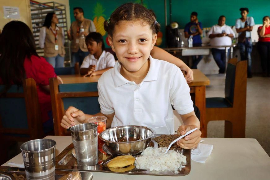 A young girl in Venezuela sits eating from a tray of food that includes rice and an arepa.
