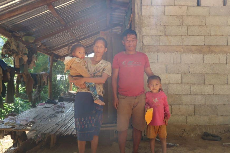 In Laos, a mother and father stand in front of a cinder block home with their two young children.