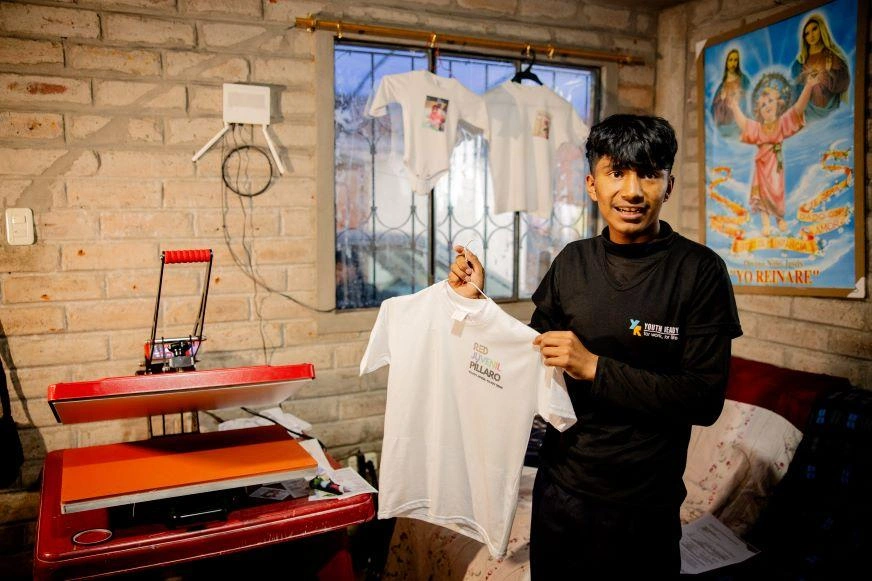 A teenage boy holds up a t-shirt beside a printing machine in Pillaro, Ecuador.