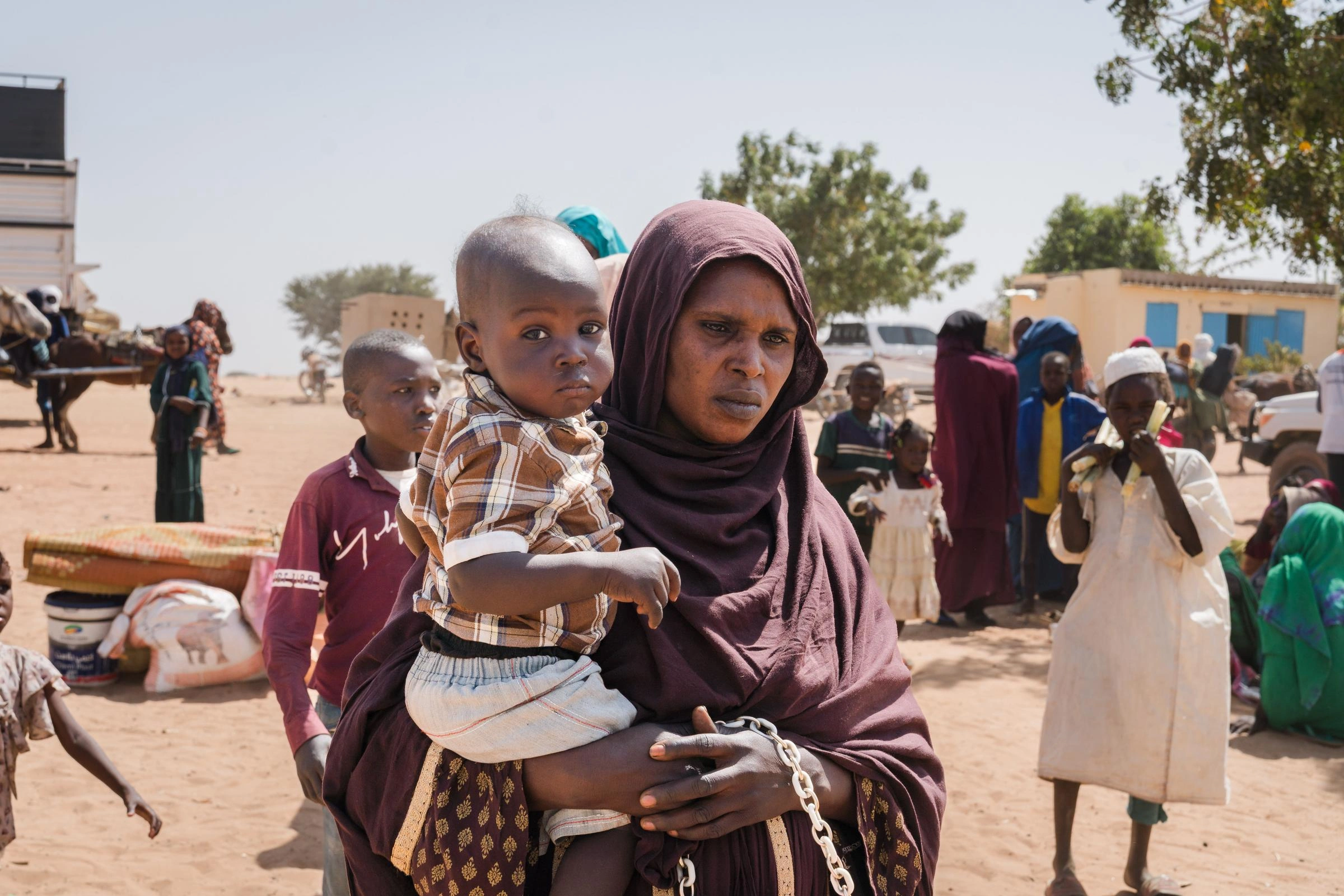 Person holding a young child in a sandy outdoor community setting, with several people, buildings, and supplies visible in the background.
