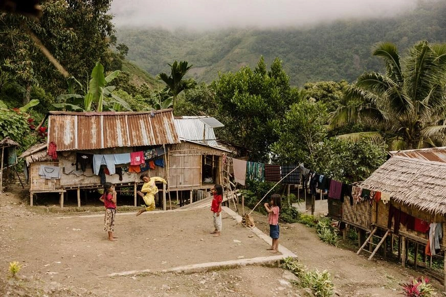 Four children play a game in a dirt yard, surrounded by wooden huts on stilts and lush vegetation in a mountainous area. (Indonesia, 2023)
