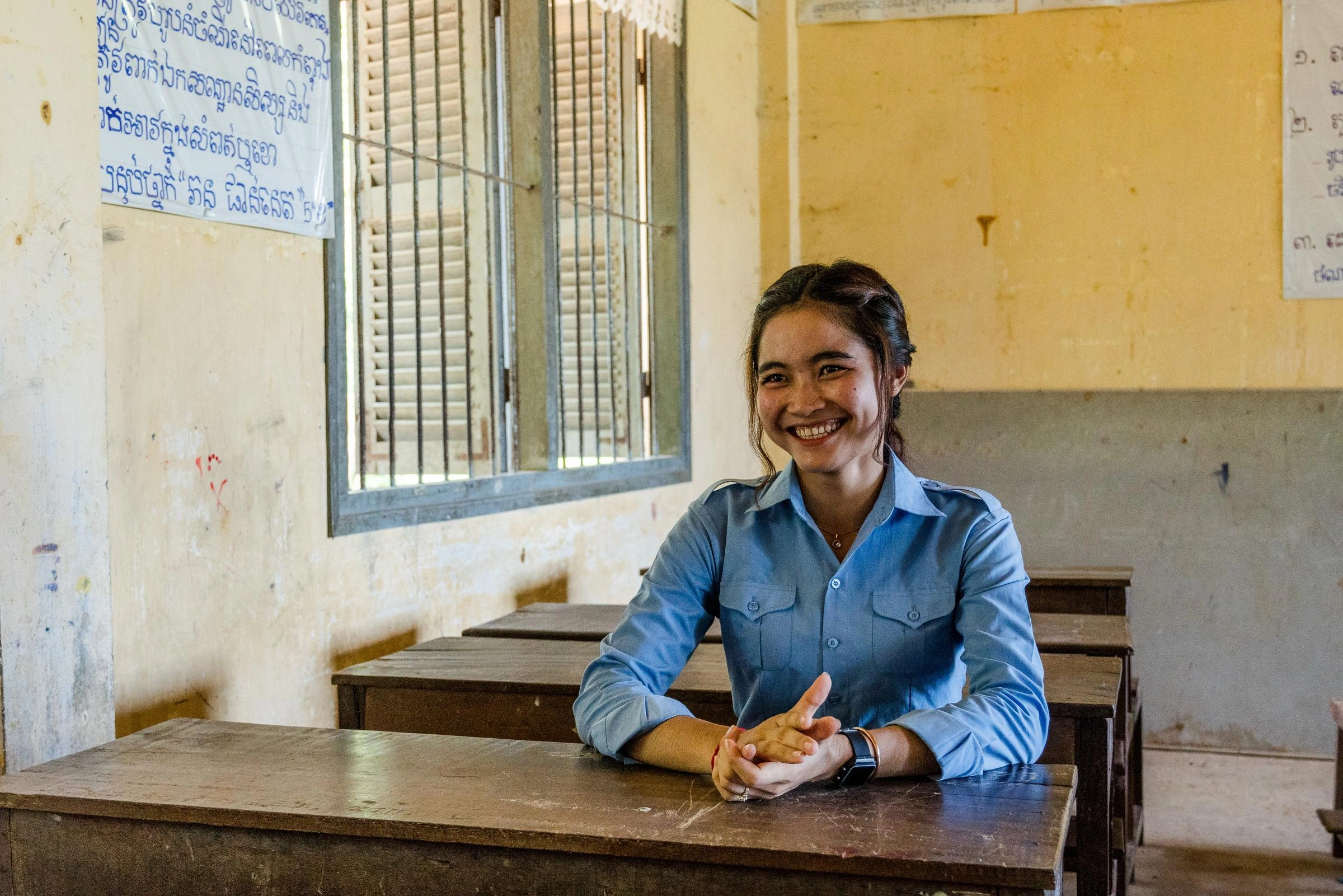 A young woman wearing a blue blouse, sits at a desk in a classroom.