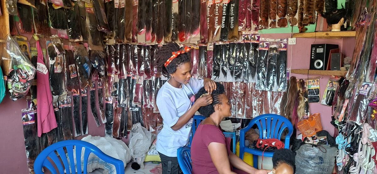 A woman sits in a blue chair as another woman in a white shirt and red headband braids her hair (Uganda, 2025)