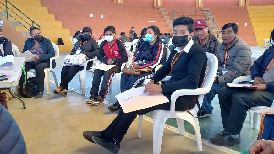 A group of teens and adults sit in white plastic chairs. Several are wearing medical masks.