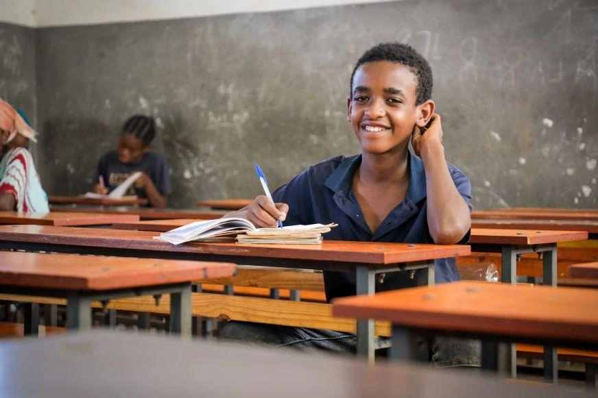 An Ethiopian adolescent boy sits at a school desk with pen and paper, smiling.