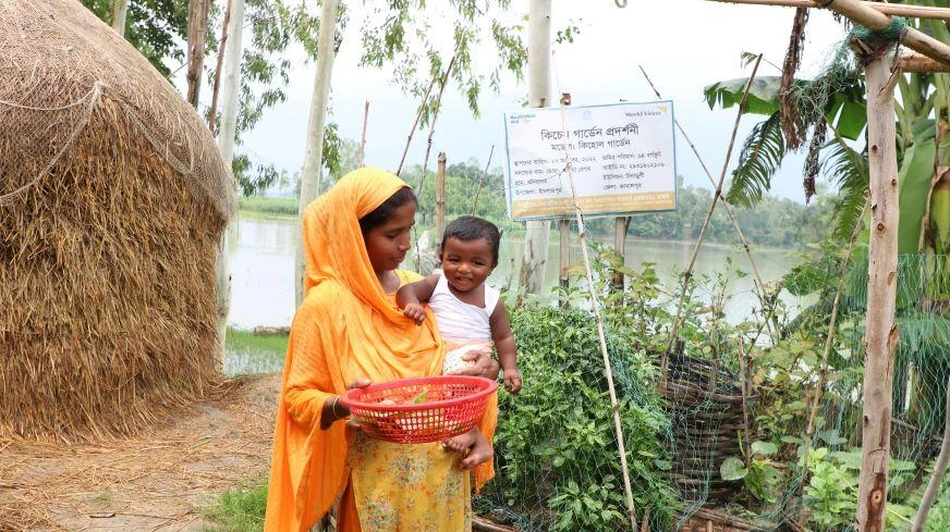 In Bangladesh, a mother dressed in orange holds her baby on her hip while collecting veggies from her garden.