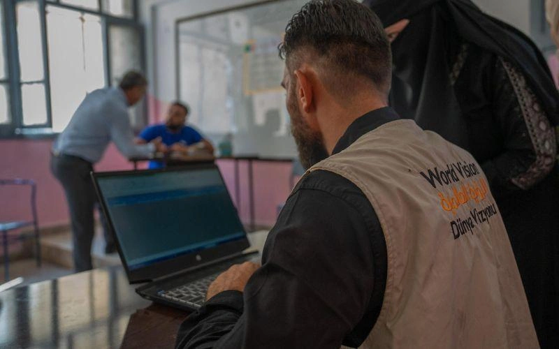 A World Vision worker types on a computer while a woman looks on at the screen, awaiting her emergency cash assistance.