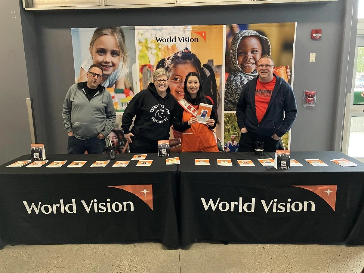Four volunteers stand behind tables branded with large World Vision logos.