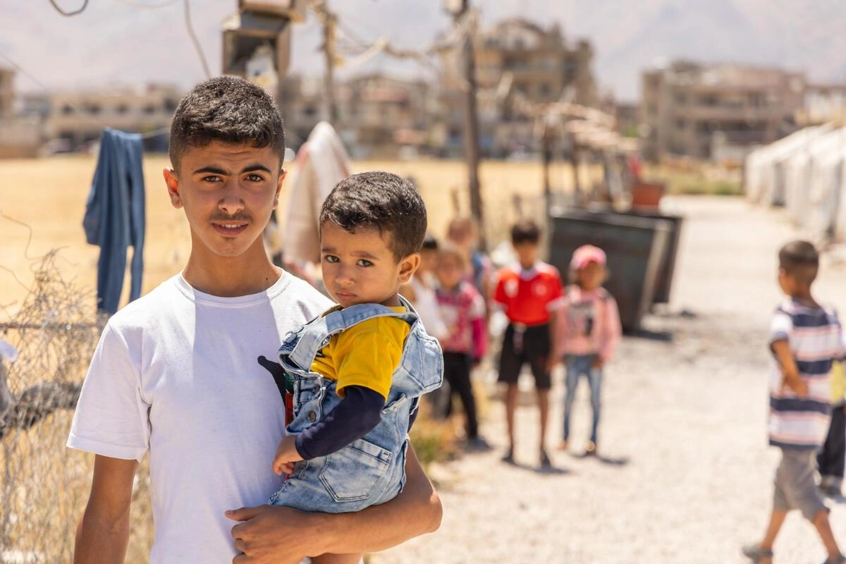 An adolescent boy holds a pre-school child, standing amidst tents.