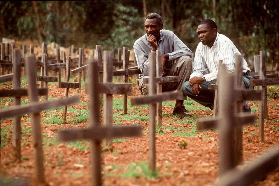 Two men squat by rows of wooden crosses.