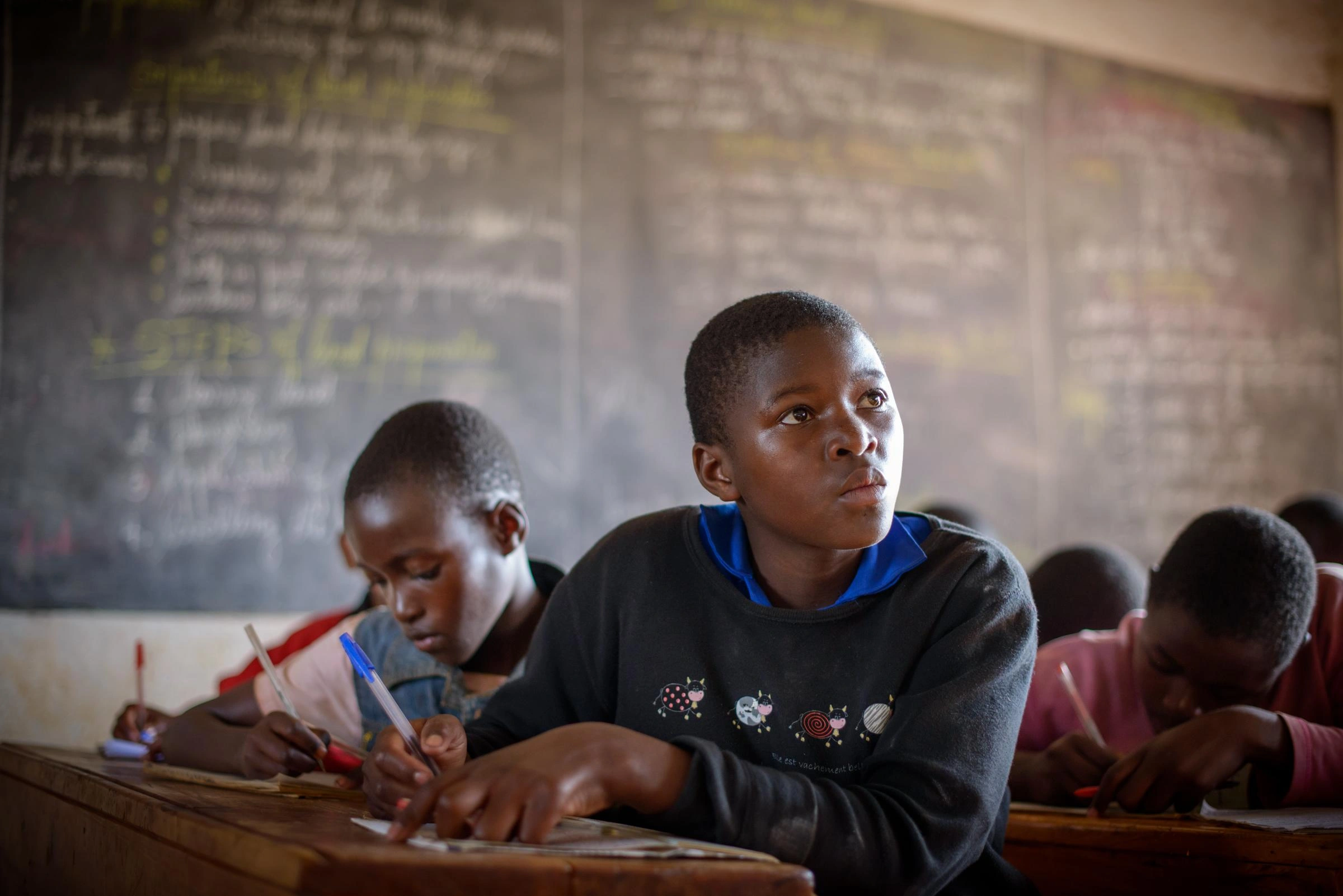 A teen girl writes on paper in a classroom full of students.