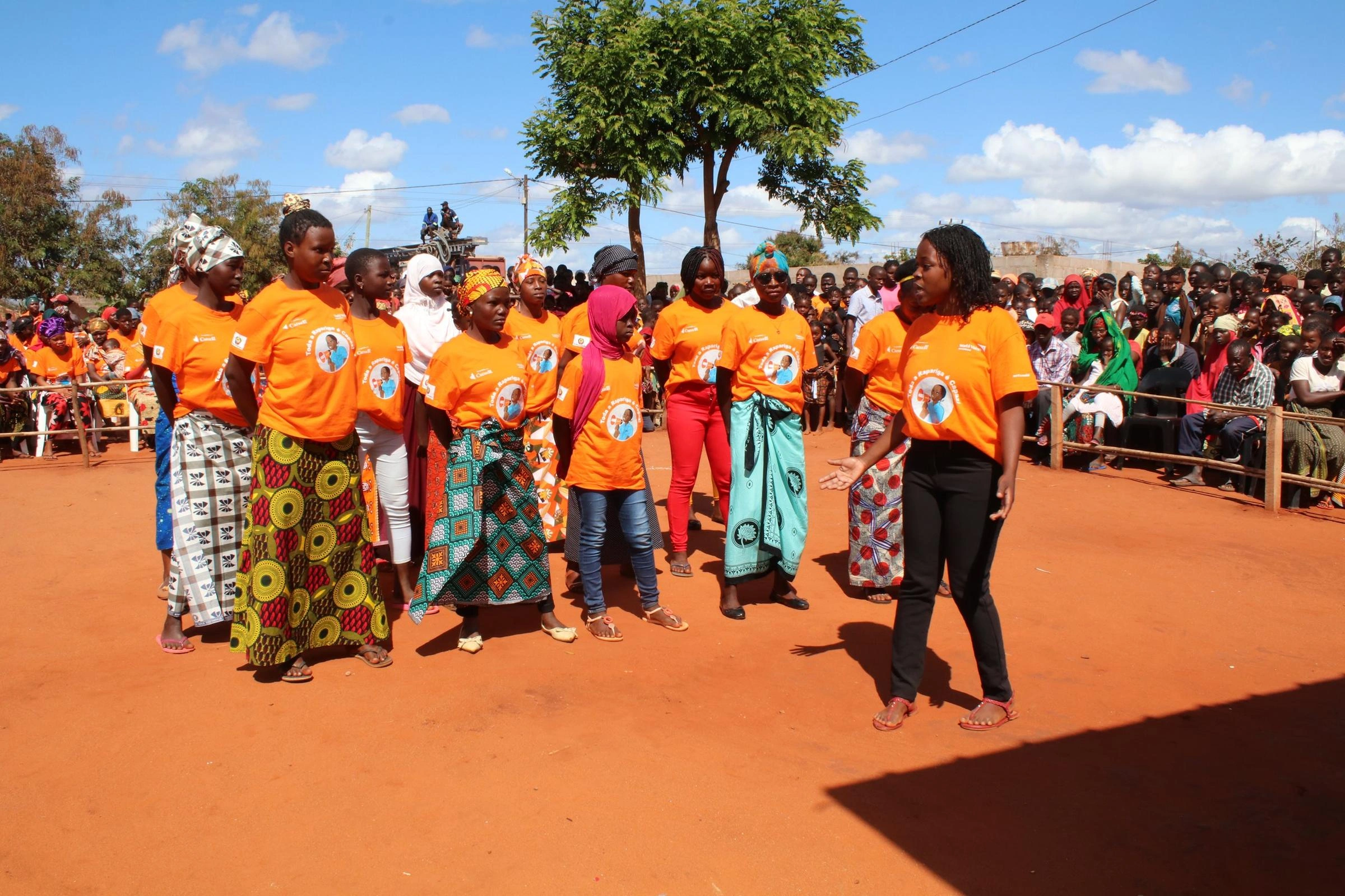 A group of girls wearing orange t-shirts, sing at a graduation ceremony.